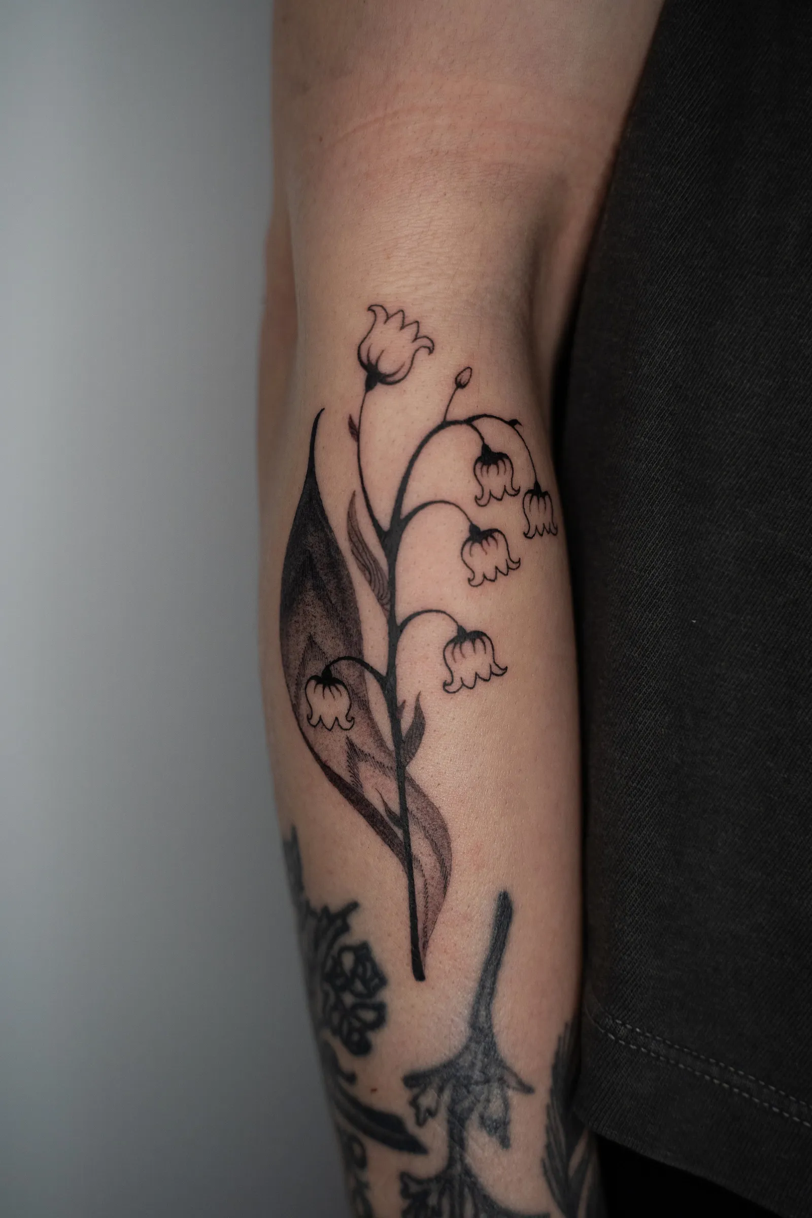 A sprig of lily of the valley, its bell flowers hanging from an arched stem beside a long leaf, fine line on a forearm